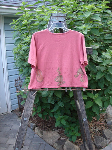 2000 Short Sleeved Crop Tee Star Music Time Coral Size 1 displayed on a wooden easel, showcasing its wide swingy shape, colorful music theme, rolled neckline, and wide short sleeves.