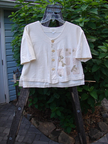 2001 Button Crop Top Pawn Natural Size 0 displayed on a wooden hanger outside, showcasing its unique vented hemline, wide short sleeves, and decorative buttons with a chess game theme.