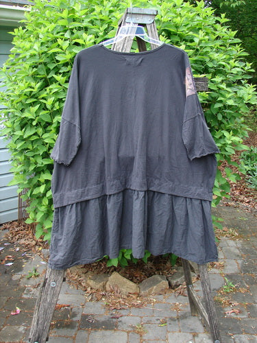 Barclay Patched Batiste Banded Flounce Esperanza Tunic Floral Storm OSFA displayed on a wooden ladder, showcasing its dolman sleeves, gathered ruffle hem, and rear birdie patch amidst leafy greenery.