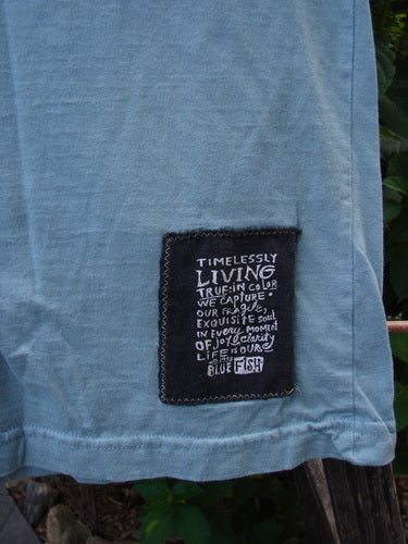 Close-up of lower hem of the 1996 Cotton Lycra Short Sleeved Layering Tee Unpainted Petal Size 2, featuring a signature black patch with poetic white text, highlighting Blue Fish's vintage charm and individuality.