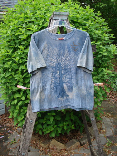 Barclay Fiona Straight Tee Tree Mottled Window Pane Size 0 displayed outdoors, featuring a large tree graphic with bird silhouettes on a blue-grey backdrop, hanging from a ladder against green shrubbery.