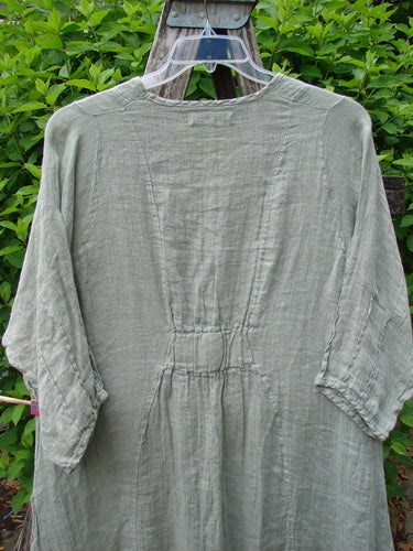 Barclay Linen High Vent Venetian Dress Contour Floral Sage Size 1, showcasing a loose fit with an oval neckline, varying hemline, and pinched three-quarter sleeves. Displayed outdoors on a hanger against leafy shrubs.