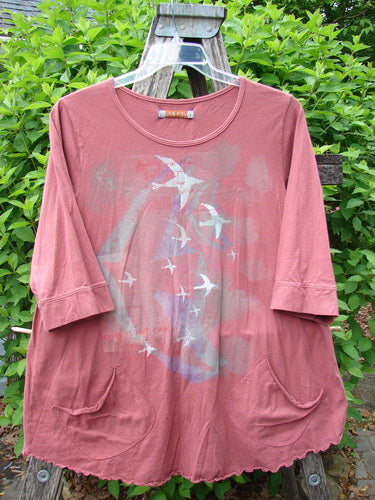 Barclay Banded Artist Pocket Top Metallic Bird Flight Rust Size 1, featuring stylized swallows, three-quarter sleeves, and ruffled hem, displayed on a wooden ladder against lush greenery.