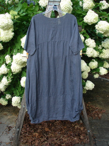 Bluefishfinder.com Barclay Linen Draw Bottom Urchin Dress in unpainted dusty navy, size 0, hangs from a wooden ladder among white hydrangeas. The linen dress has a loose, rounded silhouette, short sleeves, a gathered seam across the chest, and a softly textured finish.