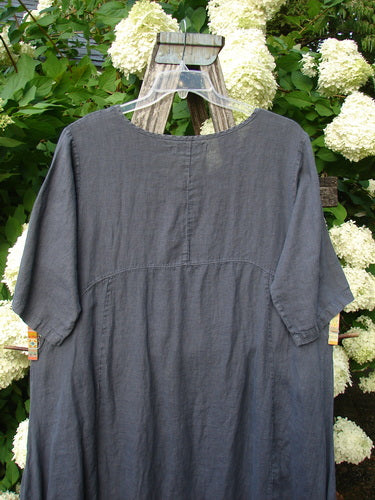 Bluefishfinder.com Barclay Linen Draw Bottom Urchin Dress in unpainted dusty navy, a loose linen dress with a round neckline, short sleeves, and softly gathered draw-bottom shaping. It hangs from a wooden ladder amid white hydrangeas, with a matte, textured drape.