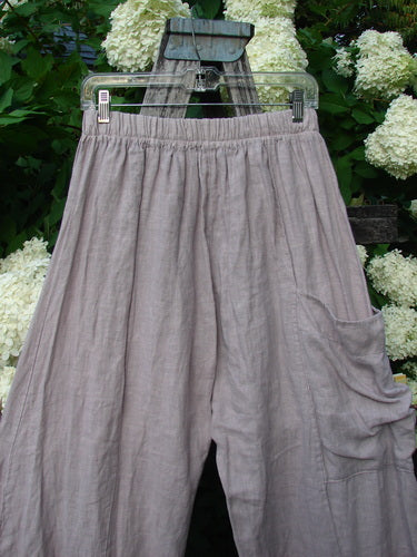 Bluefishfinder.com Barclay Linen Single Side Pocket Banded Bottom Pant in unpainted mallow, size 0, shown as loose linen pants with an elasticized banded waist and a single side pocket. The soft, lightly wrinkled fabric hangs from a clear hanger against dense white hydrangeas and dark greenery.