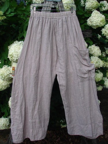 Bluefishfinder.com Barclay Linen Single Side Pocket Banded Bottom Pant in unpainted mallow, size 0, shown in a loose, crinkled linen weave with a gathered waistband, wide legs, and a single ruched side pocket. The muted gray-lilac fabric hangs against dense green foliage and pale hydrangeas.