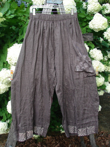 Susan Crawford Barclay Linen Single Crop Side Pocket Banded Bottom Pant in mocha, a size 2 linen pant with a side pocket and Celtic floral trim, hanging outdoors against white flowers.