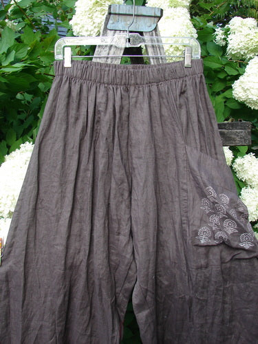 Susan Crawford Barclay Linen Single Crop Side Pocket Banded Bottom Pant Celtic Floral Mocha Size 2 in mocha linen, with a banded waist, side pocket, and floral print, hanging on a clear hanger before white flowers and green leaves.