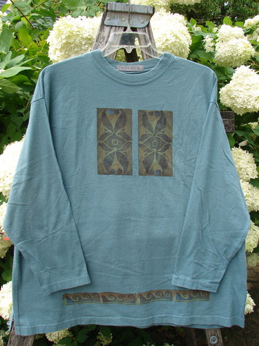 Bluefishfinder.com 1997 Long Sleeved Tee Double Celtic Flower Cypress Size 2 in faded blue-green cotton, with a crew neck, relaxed long sleeves, and a front graphic of two vertical Celtic flower panels. A narrow decorative band runs along the hem, and the shirt hangs on a hanger against white hydrangeas and green foliage.