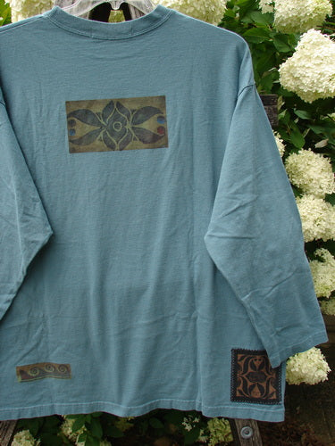 Bluefishfinder.com 1997 Long Sleeved Tee Double Celtic Flower Cypress Size 2 in faded cypress blue, a long-sleeve crewneck shirt with a soft, worn cotton look. A centered Celtic flower graphic, a small hem motif, and a square cuff print stand out against the leafy hydrangea backdrop.