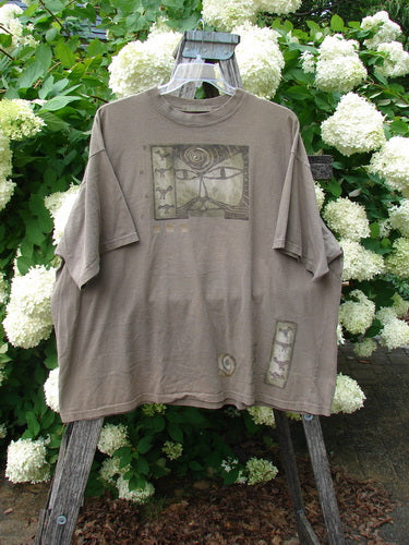 Bluefishfinder.com 1995 Short Sleeved Tee Kitty Kitty Bramble Size 2 in muted taupe, a loose short-sleeved shirt with a faded graphic panel on the chest and smaller printed accents near the hem. The soft cotton fabric hangs from a hanger outdoors, framed by dense white hydrangeas and dark green leaves.