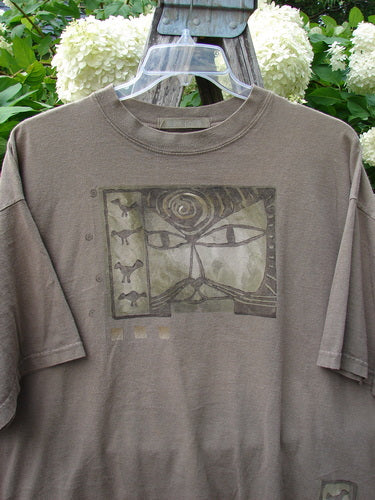 Bluefishfinder.com 1995 Short Sleeved Tee Kitty Kitty Bramble Size 2 in muted brown-gray cotton, with a faded graphic panel centered on the chest and short sleeves draping loosely from a simple crew neck. The shirt hangs on a clear hanger against leafy greenery and pale blooms.