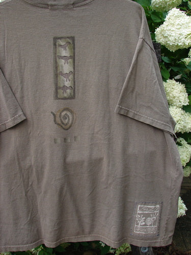 Bluefishfinder.com 1995 Short Sleeved Tee Kitty Kitty Bramble Size 2 in faded taupe cotton, with a relaxed short-sleeve cut, worn vintage texture, and a vertical graphic panel on the chest. A small printed patch sits near the lower hem, while white flowers frame the shirt at the edge of the image.