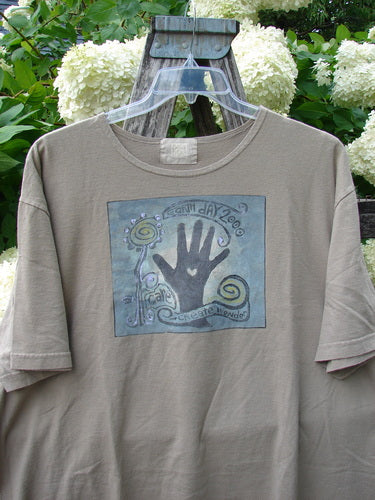 Bluefishfinder.com 2000 Artist Choice Short Sleeved Tee Earth Day Deep Dove, a muted taupe short-sleeved shirt with a centered square graphic in blue-gray tones, a black handprint, and swirling linework. The soft cotton tee hangs on a clear hanger against weathered wood and pale hydrangea blooms.