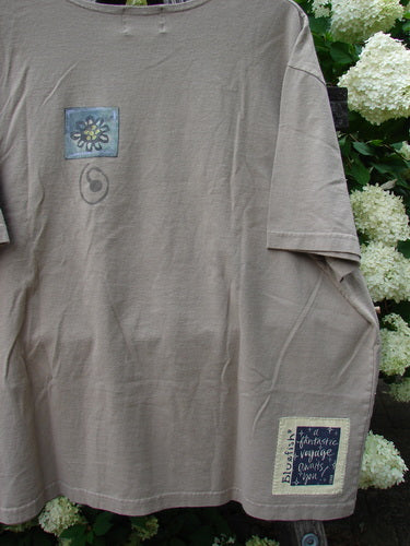 Bluefishfinder.com 2000 Artist Choice Short Sleeved Tee Earth Day Deep Dove size 2, a deep dove gray short-sleeved T-shirt with a relaxed cut, soft matte cotton texture, and visible stitched hems. Small square graphic patches sit on the chest and lower hem, hanging against white hydrangea blooms and green foliage.