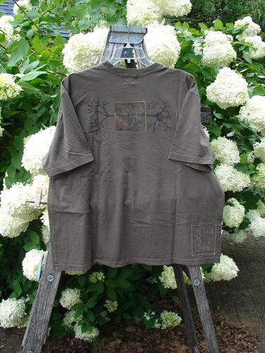 Bluefishfinder.com 2000 Short Sleeved Tee Earthen Tree Domino Size 2 in muted brown, a short-sleeved T-shirt with a faded graphic on the chest and a small square print near the hem. It hangs on a wooden ladder amid white hydrangea blooms and green foliage.