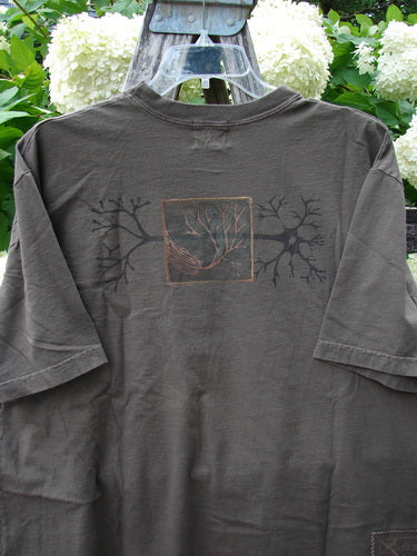 Bluefishfinder.com 2000 Short Sleeved Tee Earthen Tree Domino Size 2 in dark charcoal, a short-sleeved T-shirt with a faded tree graphic centered on the chest and a square earth-toned motif. The soft-looking cotton hangs on a hanger, with layered green foliage behind it.