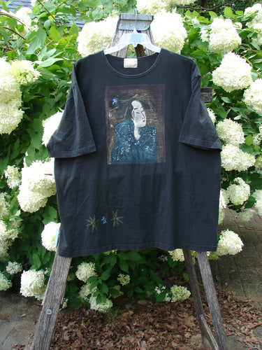 Bluefishfinder.com 1999 Short Sleeved Tee Geisha Gal Black Size 2, a faded black short-sleeve T-shirt with a centered geisha graphic in muted blue and white. The soft cotton fabric hangs loose on a wooden stand, framed by dense white hydrangeas and green foliage.