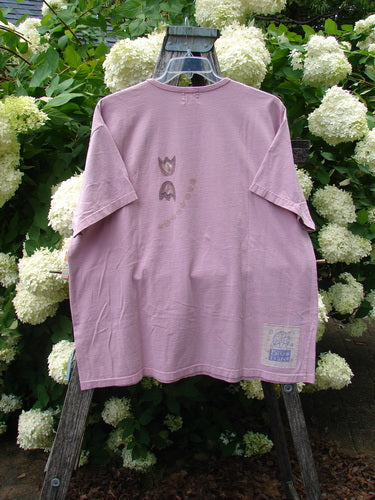 Bluefishfinder.com 1999 Short Sleeved Tee Double Daisy Peony Size 2 in faded mauve, a loose cotton T-shirt with a crew neck, short sleeves, and a soft worn finish. It hangs centered on a wooden ladder, with subtle front graphics and a small lower hem label against white hydrangeas and dense green foliage.