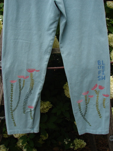 Bluefishfinder.com Barclay Cotton Lycra Crop Legging in Rising Floral Sky, size 0, shown in pale blue with slender floral stems and pink blossoms printed near the lower legs. The cropped stretch fabric hangs softly, with a stacked blue logo along one calf.
