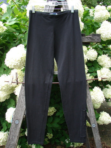 Bluefishfinder.com Barclay Cotton Lycra Side Gather Legging in unpainted black, size 0, shown as slim black leggings with a smooth stretch finish and subtle side gathering. They hang from clips on a rustic wooden frame, set against dense green leaves and white hydrangea blooms.