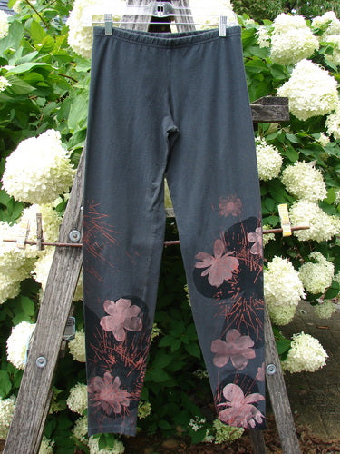 Bluefishfinder.com Barclay Cotton Lycra Legging Daisy Twig in off black, size 0, shown as slim full-length leggings with a smooth stretch knit and pink daisy-and-twig prints along the lower legs. The dark fabric hangs from a wooden ladder against white flowers and green foliage.