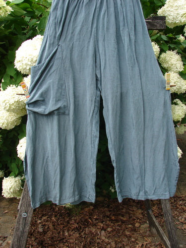 Bluefishfinder.com Barclay Silk Dimension Pant in unpainted dark sage, shown as wide, fluid silk pants with a softly wrinkled drape and loose cropped legs. The muted blue-green fabric hangs against white flowers and dark greenery, creating a layered, natural backdrop.