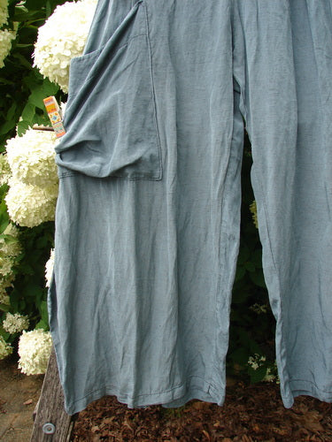 Bluefishfinder.com Barclay Silk Dimension Pant in unpainted dark sage, a pair of loose, draped silk pants with a soft matte sheen and large side pocket. The fabric falls in long vertical folds, hanging beside white flowers and dark greenery.