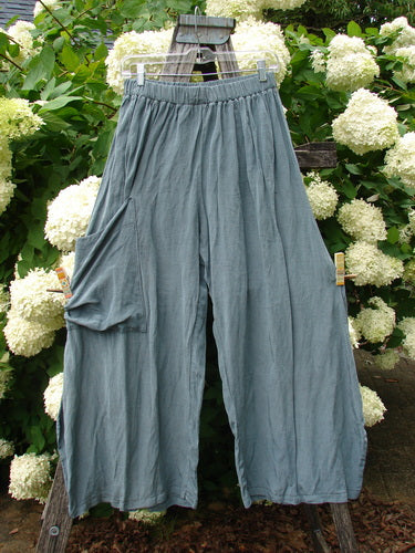 Bluefishfinder.com Barclay Silk Dimension Pant Unpainted Dark Sage Size 1, loose silk pants in a muted blue-green tone with a gathered elastic waist and wide, flowing legs. They hang from a wooden rack outdoors, the crinkled fabric catching soft daylight against dense white hydrangea blooms and green foliage.