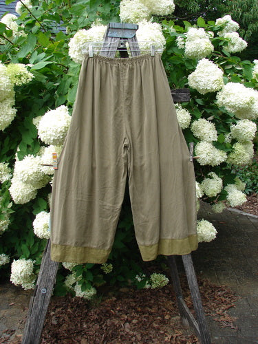 Bluefishfinder.com Barclay Lyocell Flutter Bottom Wide Leg Pant in unpainted yellow olive, size 0, hangs from a wooden frame before white hydrangeas. The soft lyocell fabric falls in a wide-leg silhouette with a fluttered hem and muted olive tone.