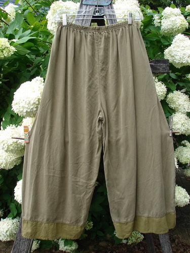 Bluefishfinder.com Barclay lyocell flutter bottom wide leg pant in unpainted yellow olive, size 0, shown in a soft drape with a high elastic waistband, wide legs, and fluttered hems. The matte fabric hangs against lush green foliage and white blossoms.