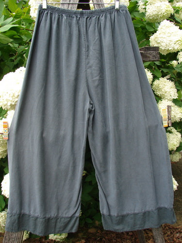 Bluefishfinder.com Barclay lyocell flutter bottom wide leg pant in unpainted light storm, shown as soft gray wide-leg trousers with a gathered elastic waist and fluttered hem. The fluid lyocell fabric drapes in a loose, airy silhouette against a leafy garden backdrop.