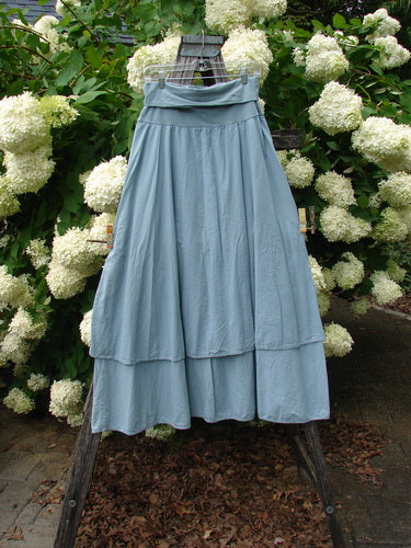 Bluefishfinder.com Barclay Batiste Fold Over Two Tier Stardust Skirt in unpainted dusky sky, a size 0 skirt with a folded-over waistband and two flowing tiers in soft, matte fabric. It hangs centered on a wooden stand, its pale blue-gray color set against dense white blossoms and green foliage.