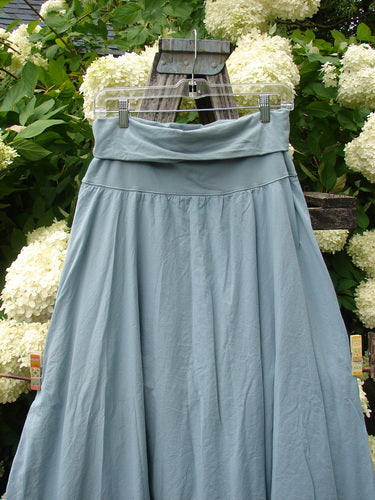 Bluefishfinder.com Barclay Batiste Fold Over Two Tier Stardust Skirt in unpainted dusky sky, a soft blue-gray skirt with a folded-over waistband and two-tier construction. It hangs from a clear hanger against leafy white hydrangeas, with a matte, lightweight drape.