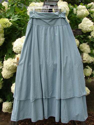 Bluefishfinder.com Barclay Batiste Fold Over Two Tier Stardust Skirt in unpainted dusky sky, a long batiste skirt with a folded waistband and two airy tiers. The soft blue fabric hangs in gentle gathers, shown against dense white hydrangeas and green foliage.