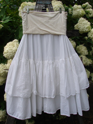 Bluefishfinder.com Barclay Linen Fold Over Two Tier Trinity Skirt in unpainted white, a layered linen skirt with a folded-over waistband, full gathered tiers, and a soft matte texture. It hangs from a clip hanger against dense green foliage and pale hydrangea blooms.