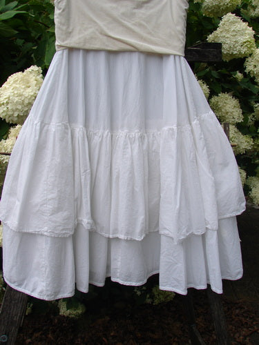 Bluefishfinder.com Barclay Linen Fold Over Two Tier Trinity Skirt in unpainted white, a layered linen skirt with a folded-over waistband and two tiered, softly gathered panels. The airy fabric falls in a loose, textured drape, framed by dark greenery and pale hydrangea blooms.