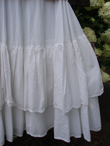 Bluefishfinder.com Barclay Linen Fold Over Two Tier Trinity Skirt in unpainted white, size 0, with layered gathered tiers and a soft linen texture. The full skirt hangs in a flowing silhouette, its pale fabric set against dark greenery.