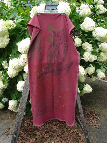 Bluefishfinder.com Barclay Heavy Weight Organic Sweatshirt Blanket Owl Forest Trimmed Burgundy OSFA hangs as a long burgundy blanket or garment, with a dark owl-and-forest graphic, trimmed edges, and a soft heavyweight fabric drape. It is displayed on a wooden stand amid white hydrangeas and green foliage.