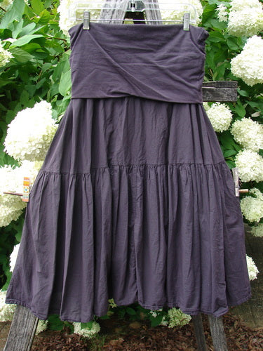 Bluefishfinder.com Barclay Linen Fold Over Single Tier Trinity Skirt in unpainted plum wine, size 0, with a folded-over waistband and a single tier of softly gathered linen that falls into a full, dark plum silhouette. The skirt hangs from clear clips against leafy white blossoms, its matte texture and layered seams visible in soft natural light.