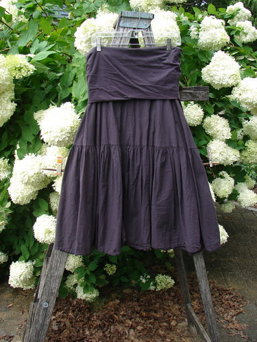 Bluefishfinder.com Barclay Linen Fold Over Single Tier Trinity Skirt in unpainted plum wine, a dark purple linen skirt with a wide fold-over waistband and a single tier of softly gathered fabric. It hangs on a rustic wooden ladder amid white hydrangeas and green leaves.
