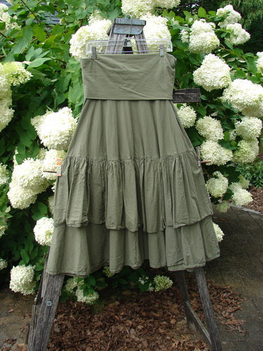Bluefishfinder.com Barclay Linen Fold Over Two Tier Trinity Skirt in unpainted dried moss, shown as a layered linen skirt with a wide fold-over waistband and two tiered ruffled panels. Its soft matte drape hangs from a rustic wooden easel amid white hydrangeas and dense green foliage.
