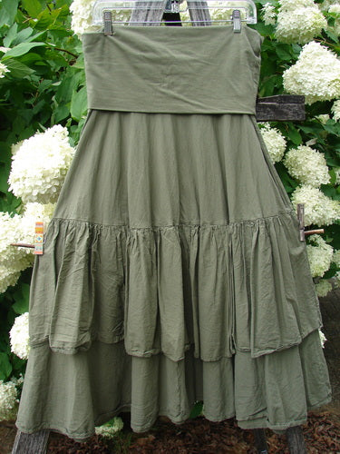 Bluefishfinder.com Barclay Linen Fold Over Two Tier Trinity Skirt in unpainted dried moss, a size 0 olive-green linen skirt with a wide fold-over waistband and layered tiered ruffles. It hangs from a clear hanger against white hydrangeas and dense greenery, with soft natural light emphasizing the matte, textured fabric.