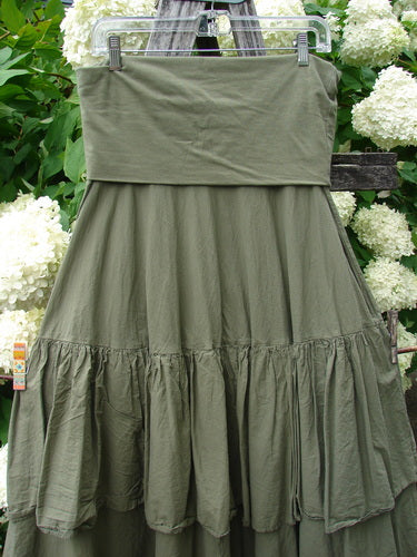 Bluefishfinder.com Barclay Linen Fold Over Two Tier Trinity Skirt in unpainted dried moss, a layered linen skirt with a wide fold-over waistband and two tiered panels. Its soft, crinkled texture and muted olive-green tone stand out against the leafy garden backdrop.