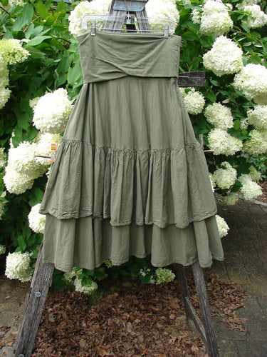 Bluefishfinder.com Barclay Linen Fold Over Two Tier Trinity Skirt in unpainted dried moss, size 0, hangs from a wooden ladder in soft olive green linen with a folded-over waistband and two tiered ruffled layers. The gathered fabric drapes against white hydrangeas and dense green foliage, creating a natural, textured outdoor display.