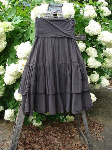 Bluefishfinder.com Barclay Linen Fold Over Two Tier Trinity Skirt Unpainted Brum Size 0 in charcoal linen, with a wide fold-over waistband and two softly tiered ruffled layers. The skirt hangs from a wooden ladder outdoors, against dense green leaves and white hydrangea blooms, under diffuse natural light.