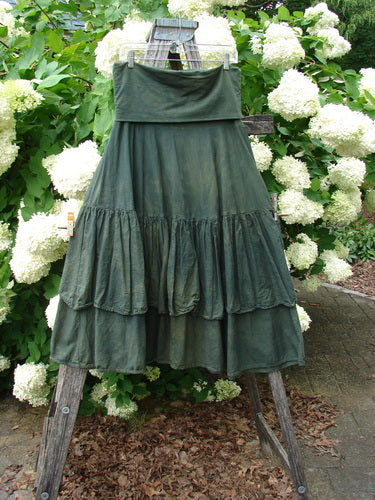 Bluefishfinder.com Barclay Linen Fold Over Two Tier Trinity Skirt in unpainted mottled army, a dark green linen skirt with a wide fold-over waistband and two tiered ruffled panels. It hangs on a wooden display stand amid white hydrangeas, with soft outdoor light and earthy tones.