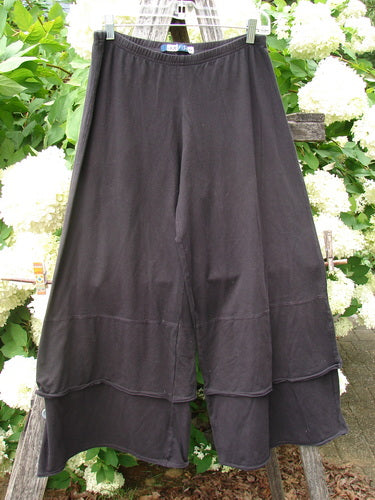 Bluefishfinder.com Barclay Cotton Lycra Crop 2 Petal Pantaloon in unpainted black, size 0, shown as a wide cropped pant with a soft stretch knit drape and layered petal hems. The dark fabric hangs in broad folds against bright green leaves and white flowers.