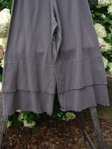 Bluefishfinder.com Barclay Cotton Lycra Crop 2 Petal Pantaloon in unpainted dusty plum, size 0, shown as wide cropped pants with a soft cotton-lycra drape and scalloped petal hems. The loose legs hang in front of leafy greenery, with muted gray-plum fabric and subtle stitched seams.